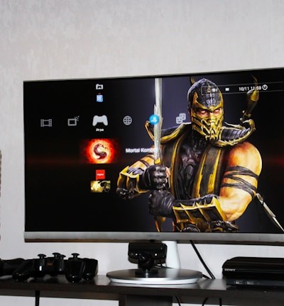 A flat-screen TV displays a video game interface featuring a character from Mortal Kombat in the center. Beside the TV is a stack of video game cases for the PlayStation 3, including titles like FIFA 14 and Grand Theft Auto. Below the TV, there is a gaming console, controllers, and other gaming equipment.