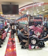 A bustling arcade filled with people playing various gaming machines. Brightly colored displays and animated characters are prominent on posters and screens. The atmosphere is lively with patrons seated in rows, focusing on the games. Overhead signage and electronic scoreboards are visible, contributing to the busy and vibrant environment.