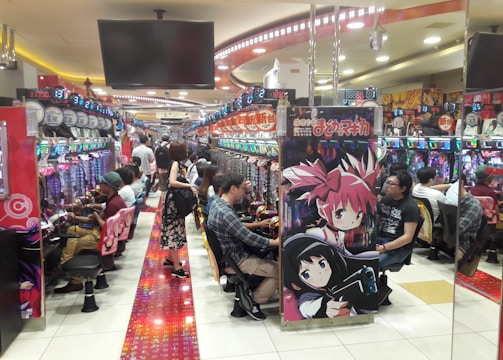 A bustling arcade filled with people playing various gaming machines. Brightly colored displays and animated characters are prominent on posters and screens. The atmosphere is lively with patrons seated in rows, focusing on the games. Overhead signage and electronic scoreboards are visible, contributing to the busy and vibrant environment.