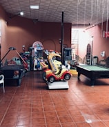 An indoor arcade area featuring various gaming machines, including an air hockey table and a ride-on toy car. The room has a tiled floor and several colorful gaming devices scattered around. Chairs and arcade signage are visible, creating a playful and entertainment-focused atmosphere.