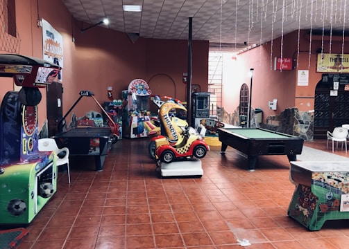 An indoor arcade area featuring various gaming machines, including an air hockey table and a ride-on toy car. The room has a tiled floor and several colorful gaming devices scattered around. Chairs and arcade signage are visible, creating a playful and entertainment-focused atmosphere.