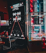 An arcade setting featuring a classic Space Invaders game setup and a modern gaming machine with seats. Bright neon lights illuminate the area, with scores displayed on digital screens. A bright and visually engaging environment full of electronic components.