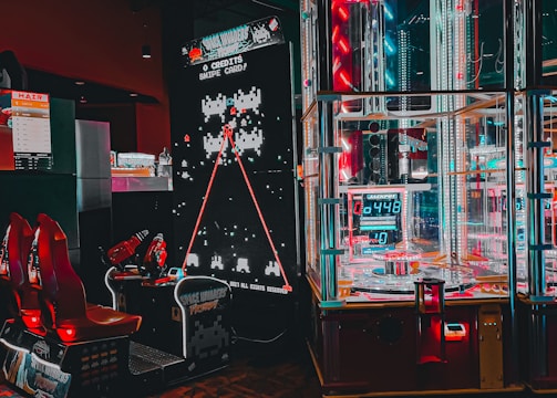 An arcade setting featuring a classic Space Invaders game setup and a modern gaming machine with seats. Bright neon lights illuminate the area, with scores displayed on digital screens. A bright and visually engaging environment full of electronic components.