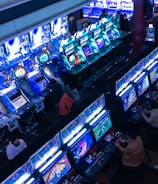 A bustling arcade filled with rows of gaming machines, each with vibrant displays and controls. Several people are seated at these machines, deeply engaged in playing games. Bright fluorescent lights cast a blue and neon glow across the room, enhancing the lively and energetic atmosphere.
