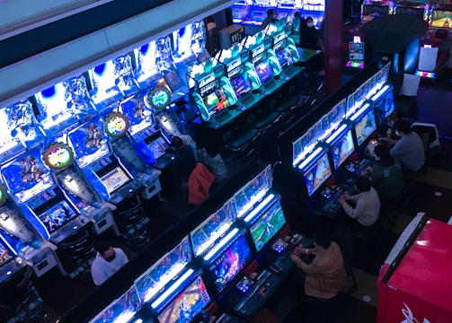 A bustling arcade filled with rows of gaming machines, each with vibrant displays and controls. Several people are seated at these machines, deeply engaged in playing games. Bright fluorescent lights cast a blue and neon glow across the room, enhancing the lively and energetic atmosphere.