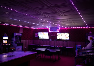 A dimly lit room with vibrant purple LED strip lights on the ceiling, featuring multiple gaming stations. There are two large screens showing video games, along with various arcade machines, including a Nintendo arcade cabinet. An air hockey table is positioned in the foreground.